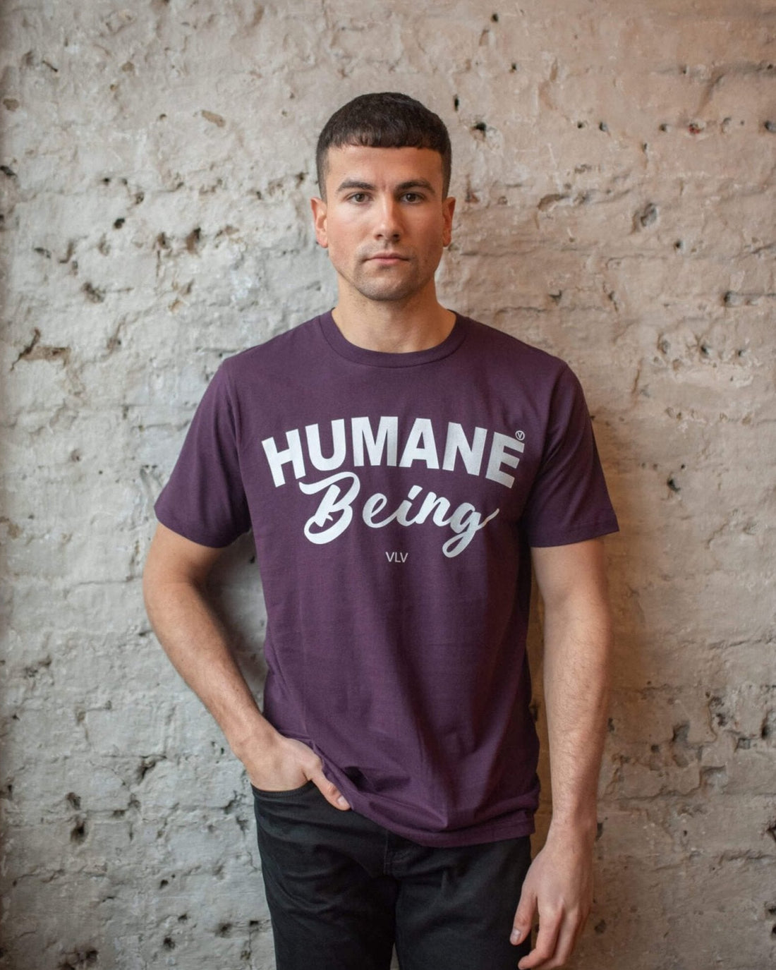 Model standing against a wall and wearing a aubergine coloured t-shirt displaying the front print of Viva La Vegan "Humane Being" eco-ethical vegan t-shirt. As worn by Russel Brand.